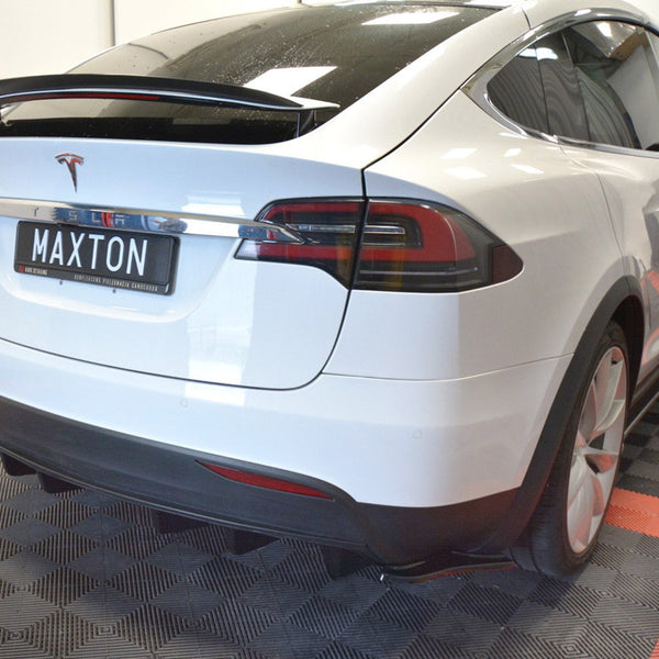 A white Tesla Model X is parked indoors on a black and red checkered floor. The license plate reads "MAXTON," and the car is positioned slightly to the right.