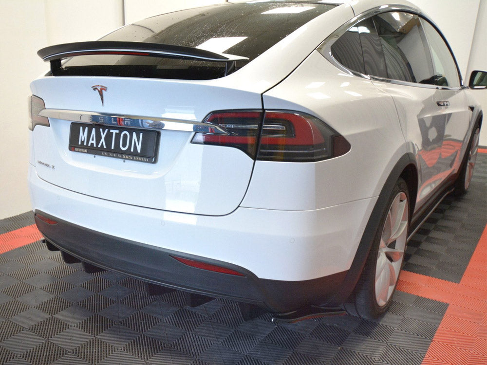 A white Tesla Model X is parked indoors on a black and red checkered floor. The license plate reads "MAXTON," and the car is positioned slightly to the right.