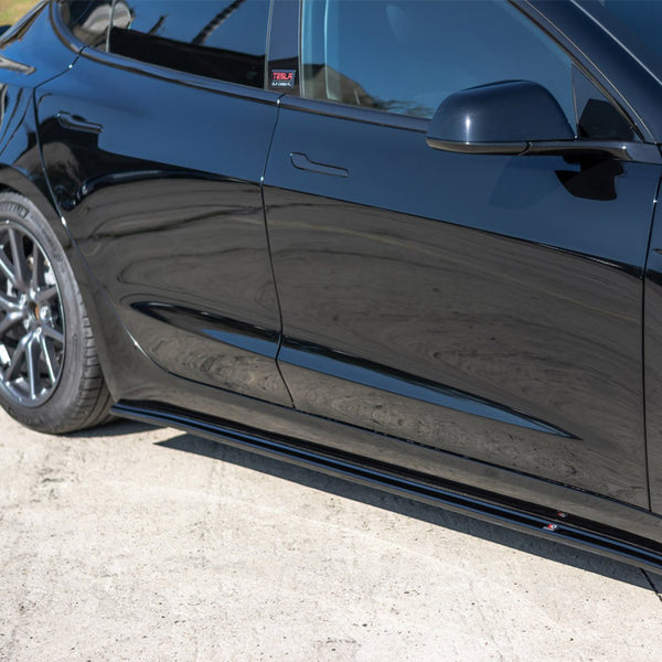 A black car is parked on a concrete surface. The side section, including the door and rear wheel, is visible. A sticker on the window reads "TESLA."