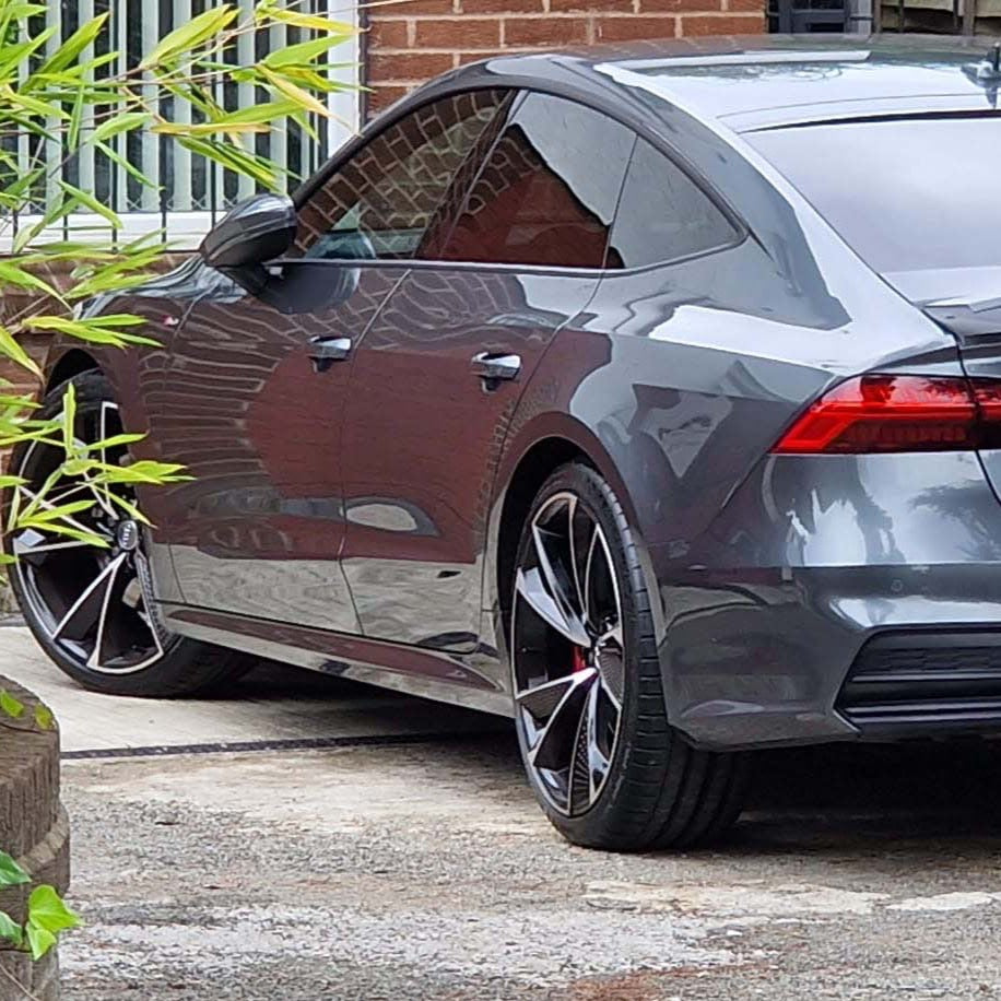 A sleek gray car is parked beside a brick building, reflecting its surroundings. Green plants partially frame the vehicle, creating a contrast between nature and architecture.