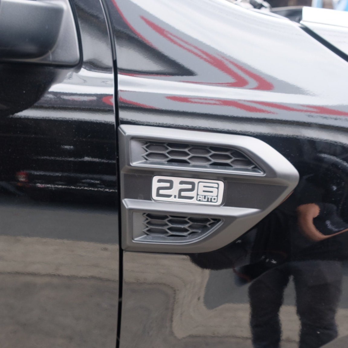 A silver vehicle emblem reads "2.2 6 AUTO" with a honeycomb design, mounted on a glossy black car surface, reflecting faint surroundings in an outdoor setting.