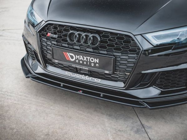 A black car's front grille displays its brand logo with a sleek design. The license plate reads "MAXTON design," and "quattro" is on the lower bumper. The car is parked on concrete.