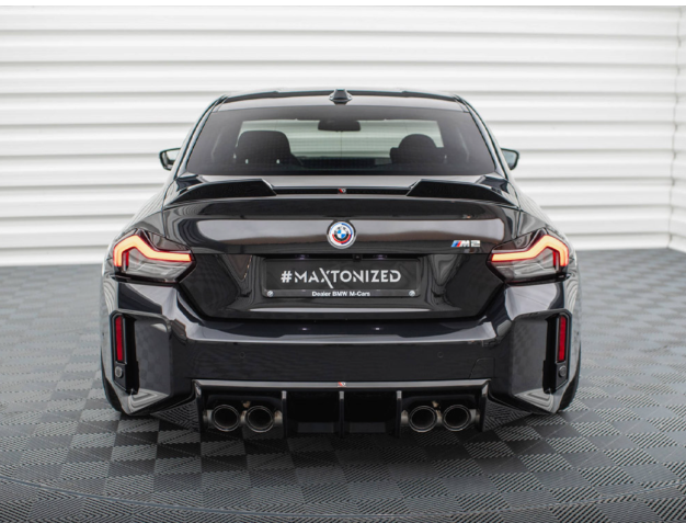 A black BMW M2 displayed in a showroom with checkered flooring. The car's rear features quad exhausts and illuminated taillights. Text on the license plate reads "#MAXTONIZED Dealer BMW M Cars."