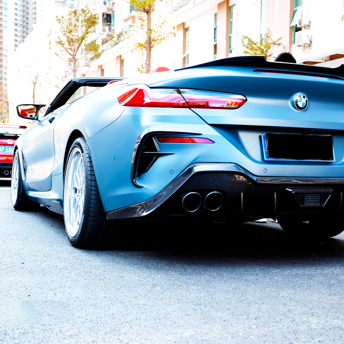 A blue convertible BMW, parked on a city street, is viewed from behind, with red cars lined up ahead. Buildings and trees provide an urban backdrop.