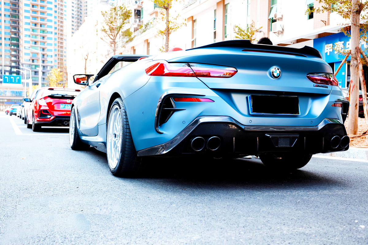 A blue convertible BMW, parked on a city street, is viewed from behind, with red cars lined up ahead. Buildings and trees provide an urban backdrop.