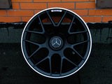 A black AMG car wheel rests against an orange brick wall on asphalt. The wheel features a prominent silver Mercedes emblem at the center.