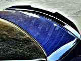 A shiny black car's trunk features a sleek aerodynamic spoiler. It reflects surrounding trees and sky, parked on a textured concrete surface in a well-lit area.