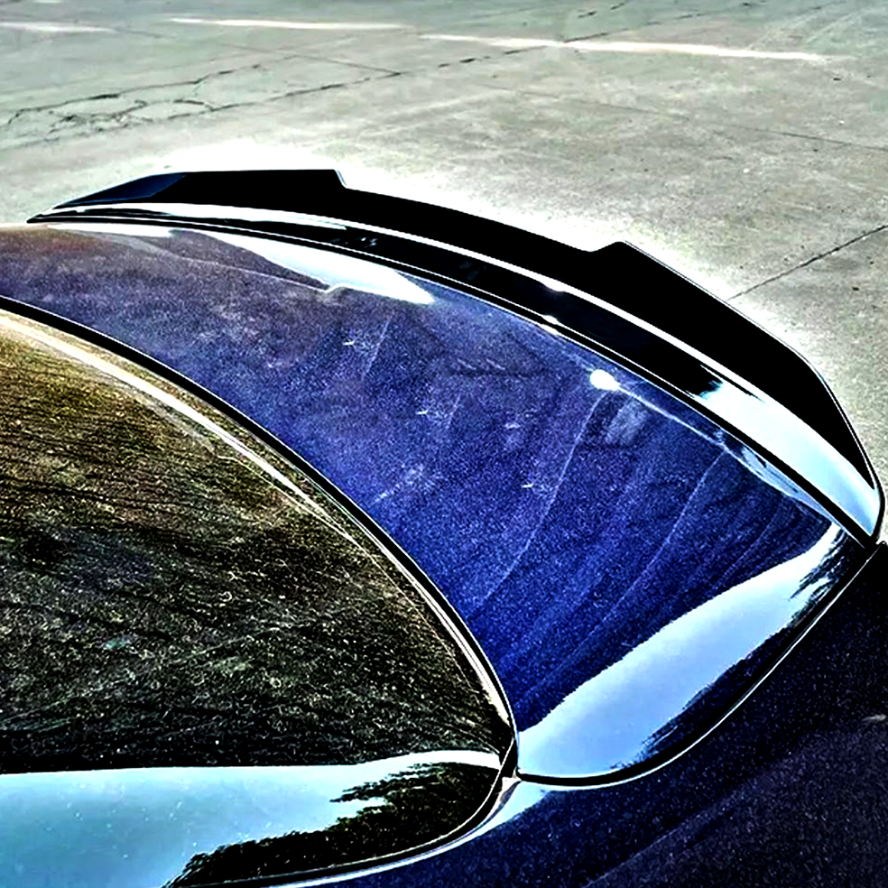 A shiny black car's trunk features a sleek aerodynamic spoiler. It reflects surrounding trees and sky, parked on a textured concrete surface in a well-lit area.