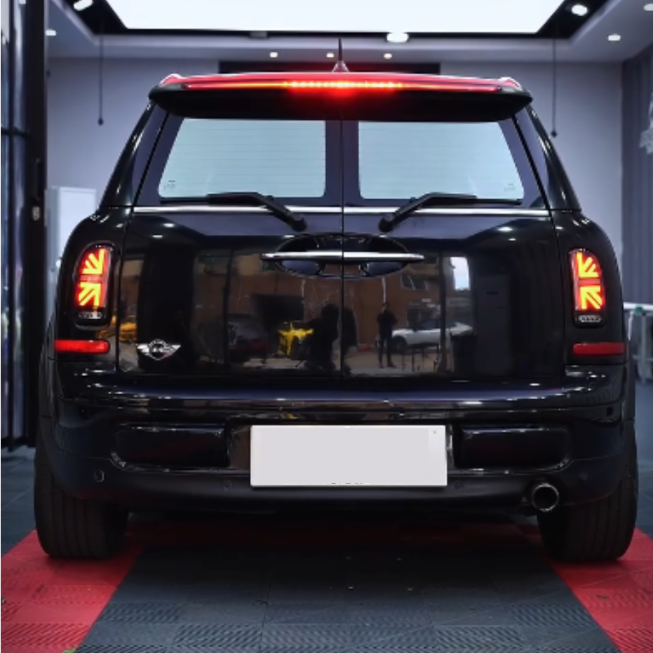 A black car is stationary on a red and black checkered garage floor. The car's rear lights are illuminated, with the surrounding area reflecting in its glossy surface.