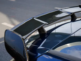 A carbon fiber car spoiler sits on a blue vehicle, reflecting the sky. It's positioned beside a road with a concrete barrier, suggesting a street-setting.