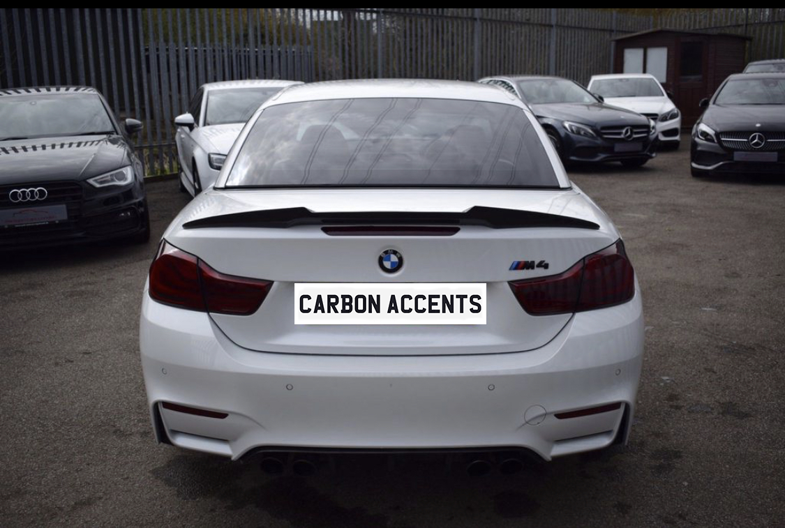 White BMW M4 parked with visible rear carbon accents, in a car lot surrounded by multiple vehicles. License plate reads "CARBON ACCENTS."
