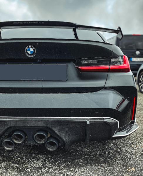 The rear view of a black BMW car with a visible logo, quad exhausts, a "M Performance" spoiler, and red taillights, is parked on gravel with cloudy skies overhead.