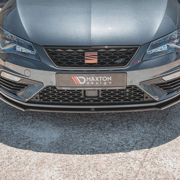 A gray car's front view features sharp headlights and a honeycomb grille. It displays a "M✪AXTON design" license plate on concrete pavement in bright sunlight.