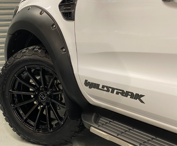 A white vehicle features black fender flares and off-road tires. Text on the side reads "WILDTRAK" and "BI-TURBO." The backdrop appears to be a garage with a gray metallic door.
