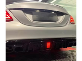 A white car's rear features illuminated taillights and a glowing red light on the diffuser. It's parked indoors on a patterned floor, with soft overhead lighting.