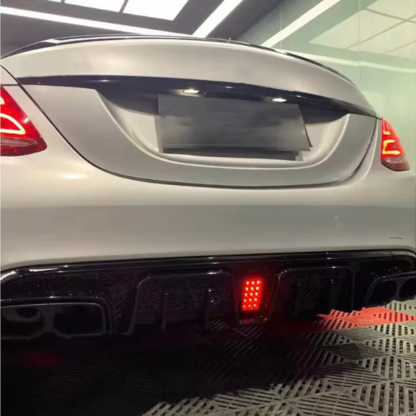 A white car's rear features illuminated taillights and a glowing red light on the diffuser. It's parked indoors on a patterned floor, with soft overhead lighting.