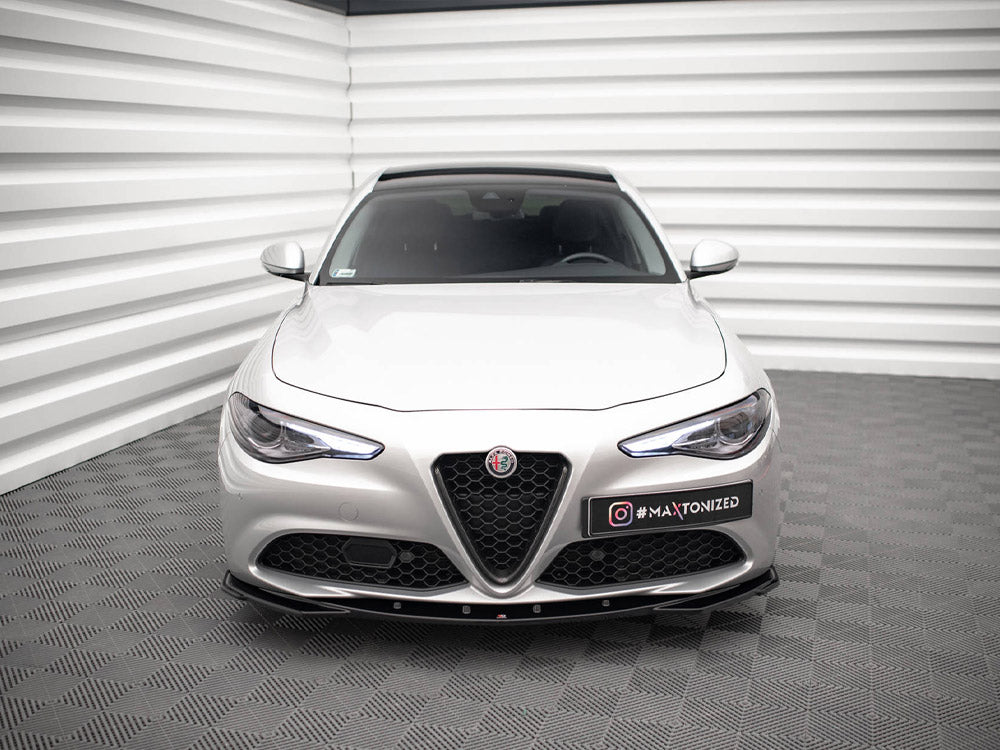 A silver car is parked in a modern showroom with textured walls and flooring. The car's front features a distinctive emblem and a license plate reading "#MAXTONIZED."