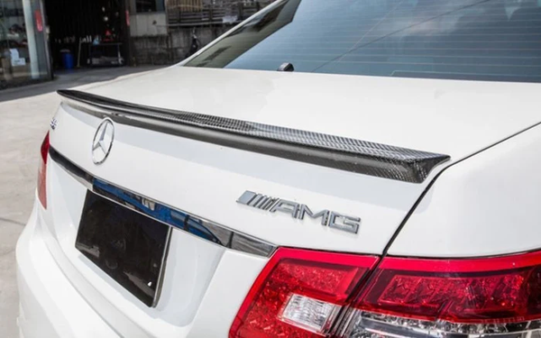 A white car with AMG and Mercedes logos features a carbon fiber spoiler on the trunk lid, parked near a building with various objects in the background.
