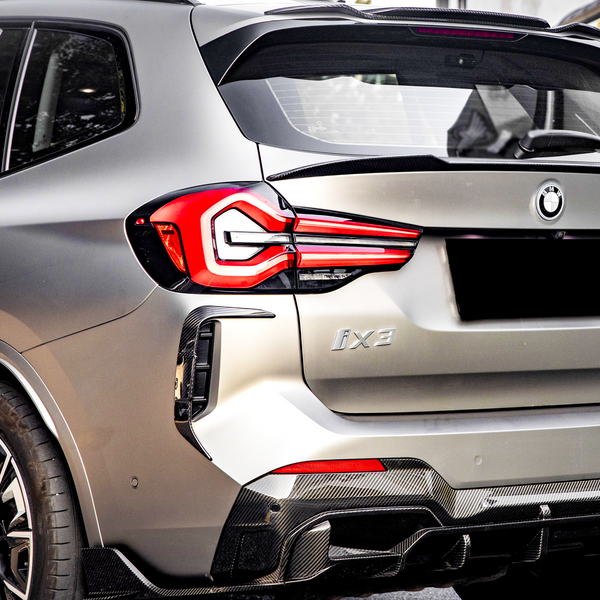 A metallic gray SUV with prominent red tail lights is parked outdoors. The rear badge reads "iX3," and features a BMW logo, indicating its model in a bright setting.