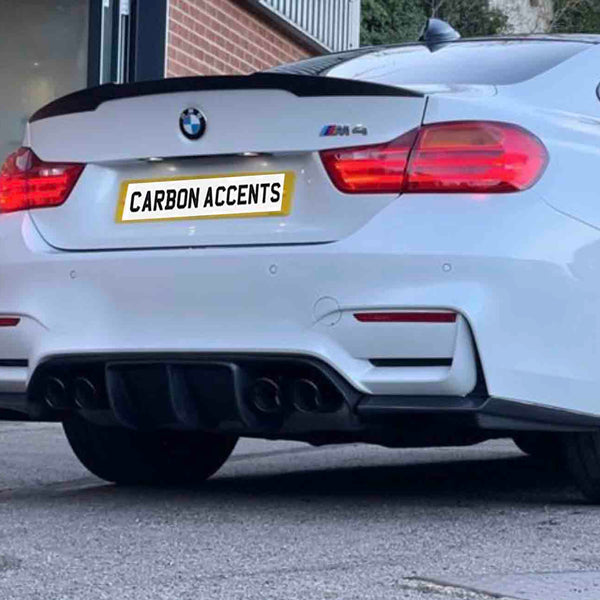 A white BMW M4 is parked outside a modern building. The car's license plate reads "CARBON ACCENTS." The rear features visible exhausts and red tail lights.