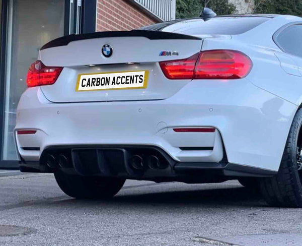 A white BMW M4 is parked outside a modern building. The car's license plate reads "CARBON ACCENTS." The rear features visible exhausts and red tail lights.