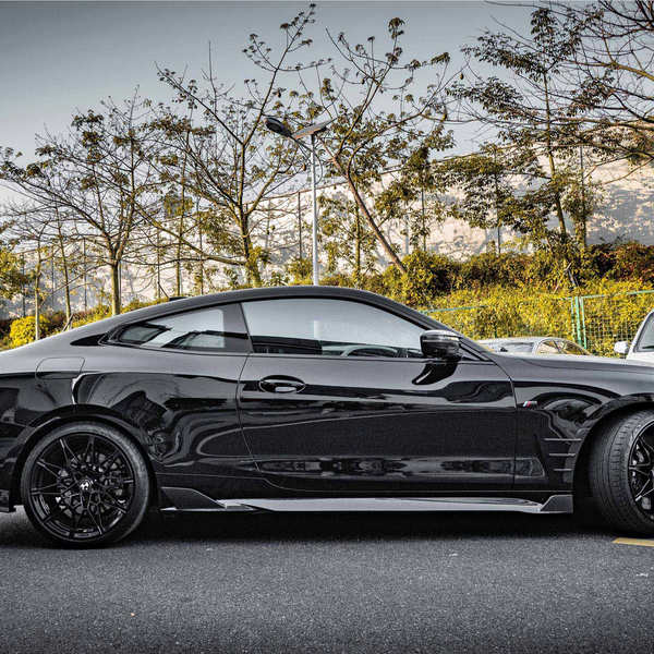A sleek black sports car is parked on a roadside. Trees with sparse foliage line the background, alongside other parked vehicles under a clear sky.
