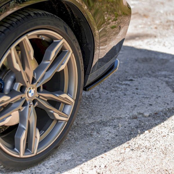 A car tire with a distinctive alloy wheel design is stationary on a rough, sunlit gravel surface, partially casting a shadow. The BMW logo is visible on the hubcap.