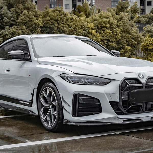 A sleek white BMW is parked on wet pavement, featured prominently in a spacious outdoor parking lot, bordered by lush green trees. Text found: "M Performance."