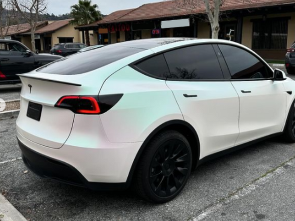 A white car with tinted windows and black wheels is parked in a lot. Surroundings include other vehicles and buildings with signs such as "TATTOO" visible.