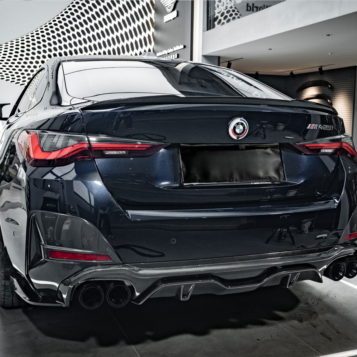 A sleek, dark-colored car is parked in a showroom with a modern design. The car's rear features prominent taillights and quad exhaust pipes. The wall has text: "wrkz DryCarbon™."