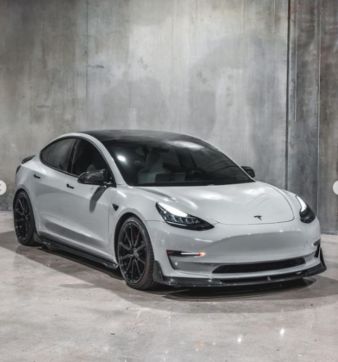 A white Tesla car is parked in a concrete-walled room, displaying a sleek, modern design with black-rimmed wheels and tinted windows.