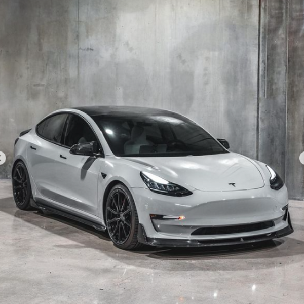 A white Tesla car is parked in a concrete-walled room, displaying a sleek, modern design with black-rimmed wheels and tinted windows.
