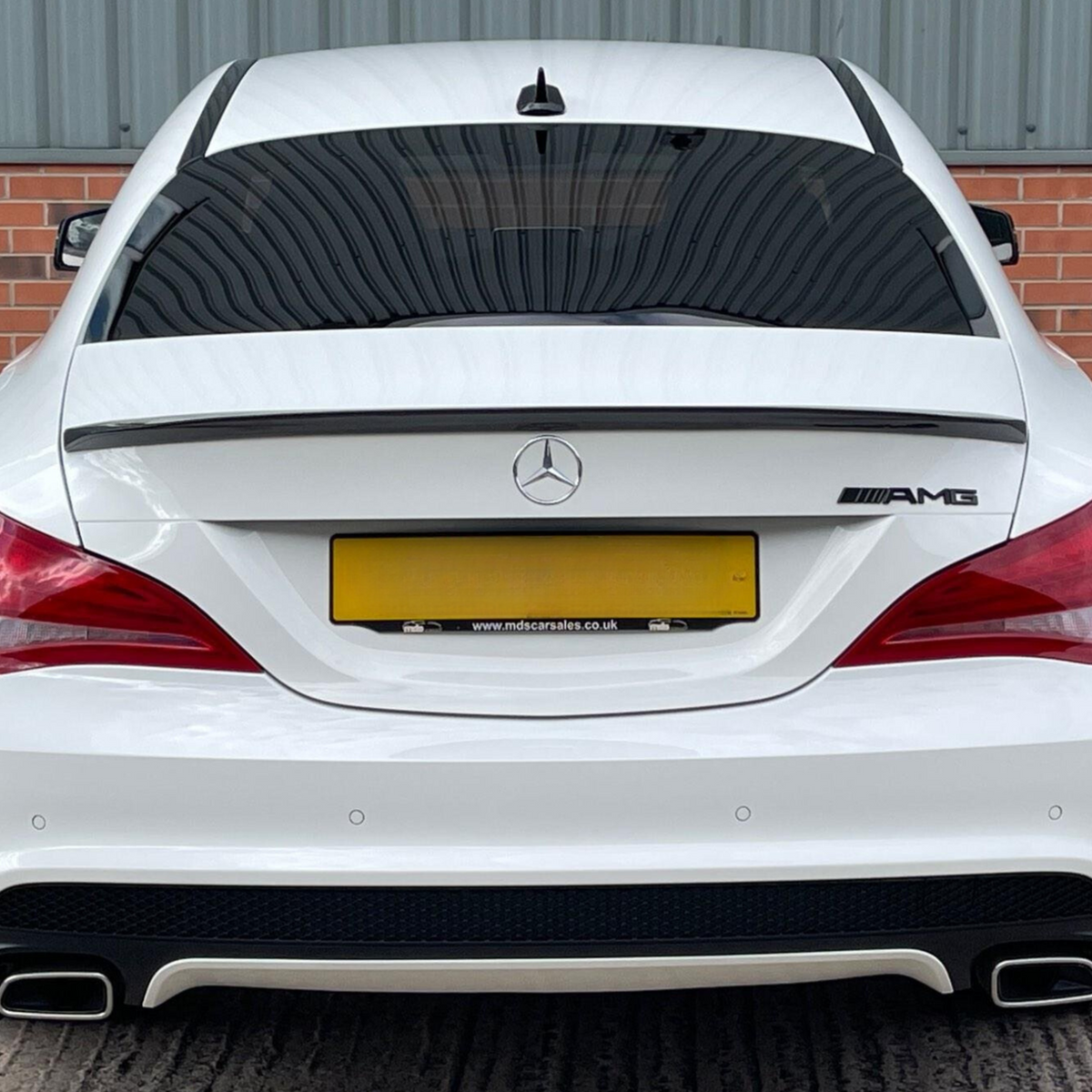 A white Mercedes-Benz AMG car is parked, featuring dual exhausts and tinted windows, against a backdrop of red brick and corrugated metal. Text: "www.mdscarsales.co.uk".