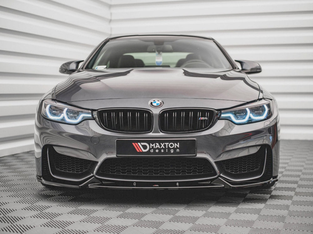A sleek, dark BMW with illuminated headlights sits on a patterned floor, facing forward. Behind it is a textured, white wall. The front license plate reads "MAXTON design."