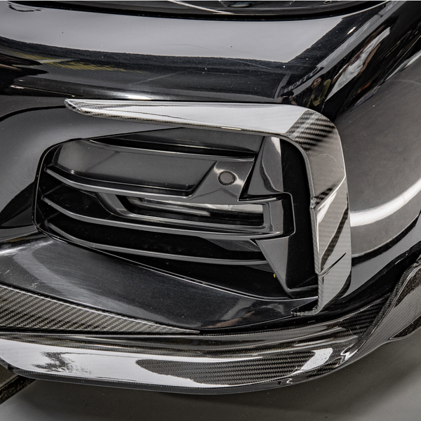 Close-up of a car’s front bumper featuring a sleek, black grille with carbon fiber trim detailing; the reflection and lighting highlight the material's texture in an indoor setting.