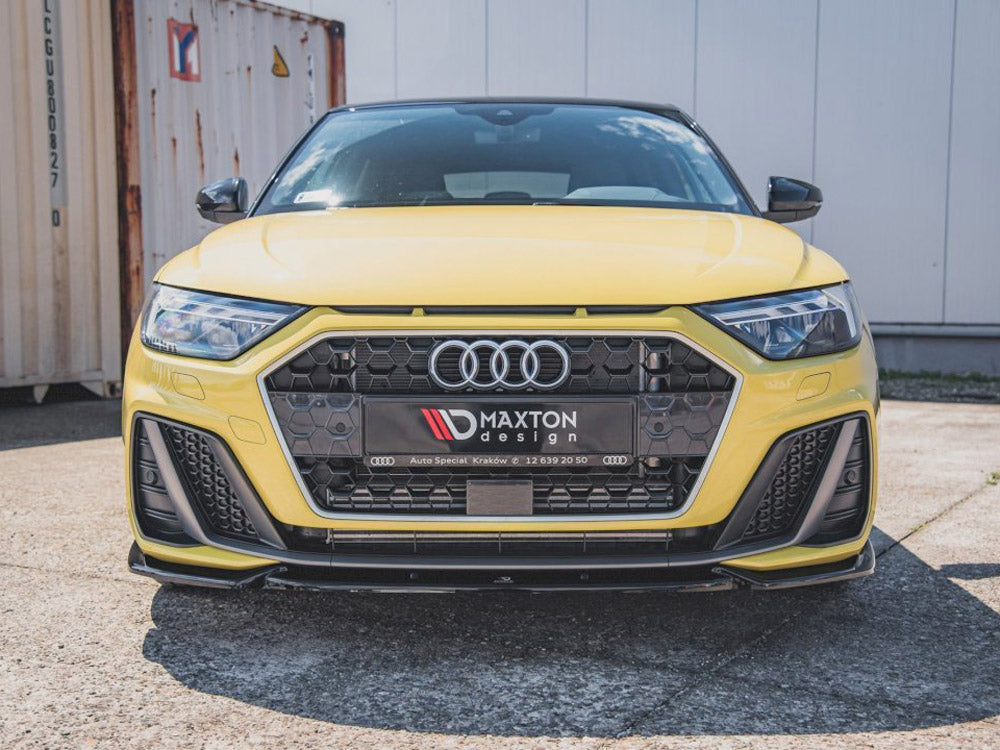 A yellow Audi car is parked on pavement, featuring a prominent hexagonal grille with Maxton design branding. Shipping containers and a white building are visible in the background.