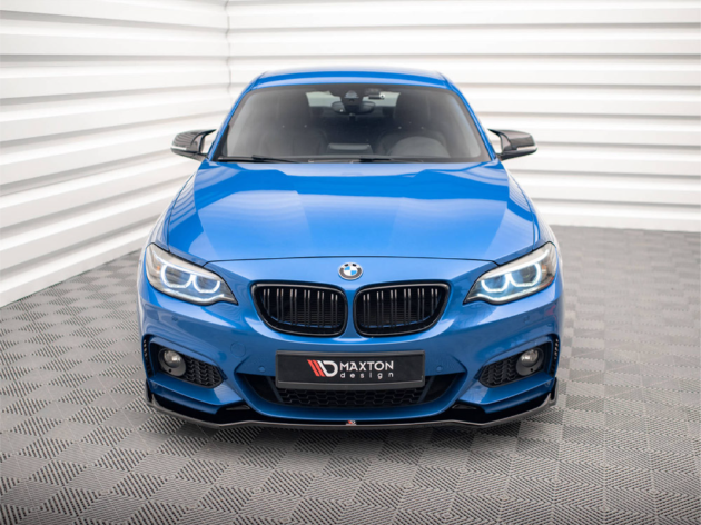A blue BMW car, parked on a patterned gray floor, faces forward. Bright headlights illuminate, surrounded by white, ribbed walls. A sign reads "MAXTON design" on the front.