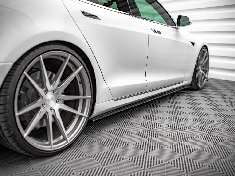 A sleek white car, viewed from the side, is parked on a geometric-patterned floor in a modern, white-paneled room. The rims feature a large, angular design.