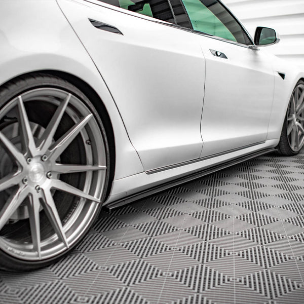 A sleek white car, viewed from the side, is parked on a geometric-patterned floor in a modern, white-paneled room. The rims feature a large, angular design.