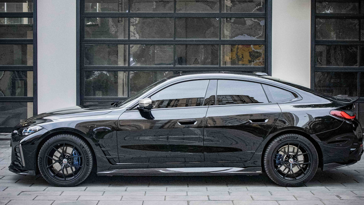 A sleek black car is parked on a cobblestone surface, reflecting nearby buildings, in front of a modern building with large, grid-like windows.