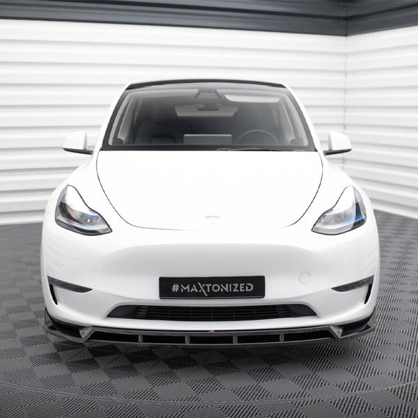 A white car is parked in a corner of a room with white and gray walls and flooring. The license plate reads "#MAXTONIZED."