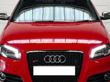 A red Audi car, stationary, with headlights on, is parked indoors against a white brick wall. The car displays a black grille with the Audi logo and a blank license plate.