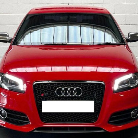 A red Audi car, stationary, with headlights on, is parked indoors against a white brick wall. The car displays a black grille with the Audi logo and a blank license plate.
