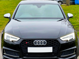 A black Audi car is parked on a road, facing forward. It features distinctive headlights and a front grille with the Audi logo. In the background, grassy areas and other parked cars are visible.