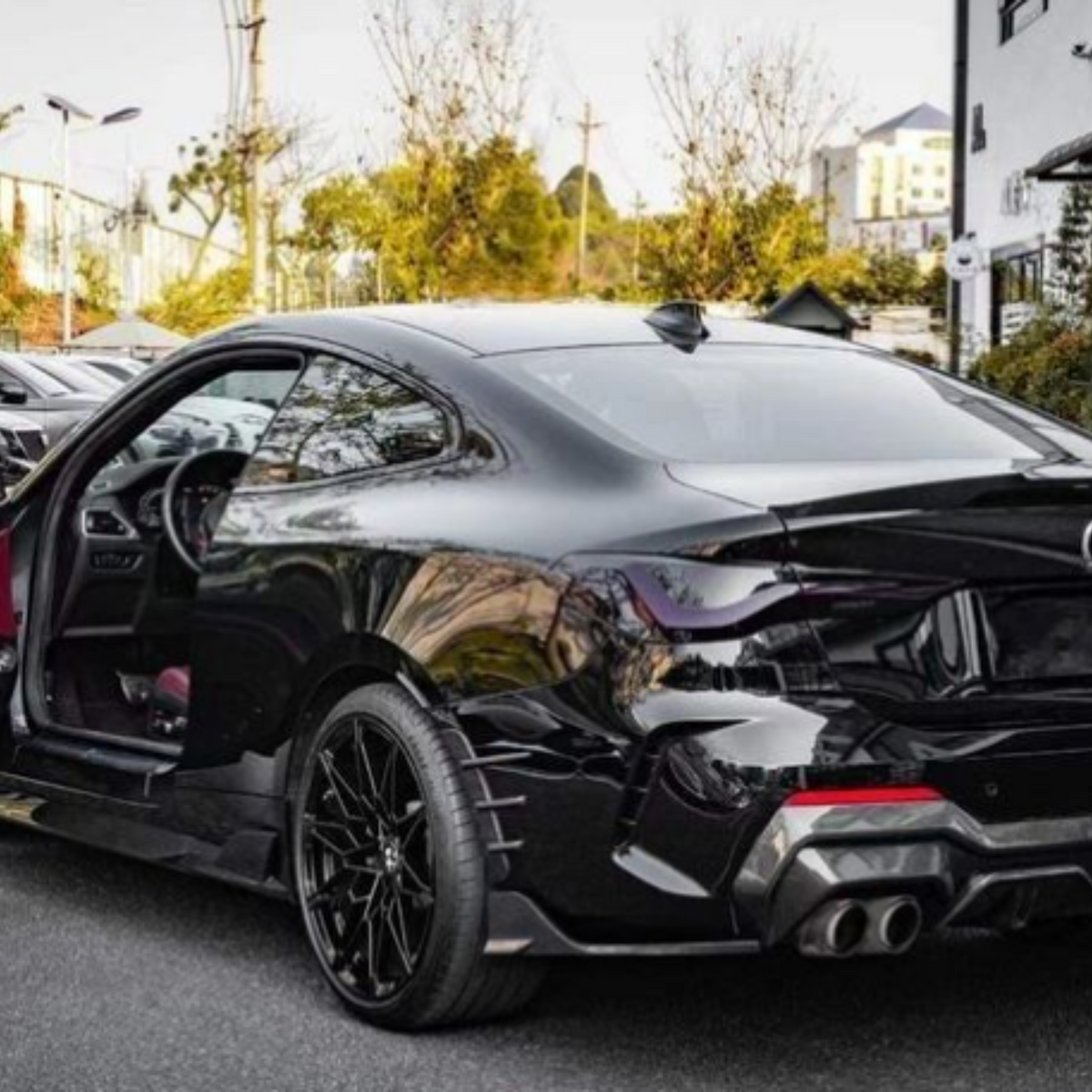 A sleek black car with an open driver's door revealing a red interior is parked on a street, surrounded by trees and modern buildings.