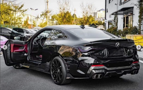 A sleek black car with an open driver's door revealing a red interior is parked on a street, surrounded by trees and modern buildings.