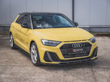 A yellow Audi car is parked on a concrete surface. The front grille features the Audi logo. Text on the grille reads 
