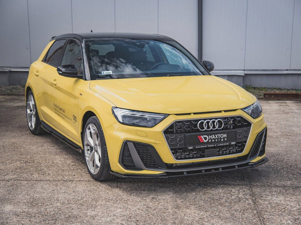 A yellow Audi car is parked on a concrete surface. The front grille features the Audi logo. Text on the grille reads "MAXTON design." A gray industrial building is in the background.