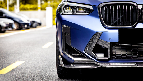 A close-up of a blue car's front detailing its grill and headlights, parked on a road lined with yellow markings, with blurred cars visible in the background.