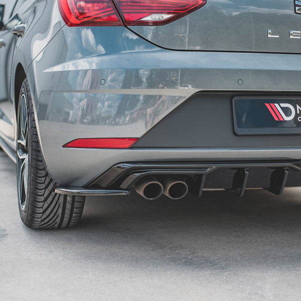 The rear of a gray car, featuring dual exhaust pipes. "LEON" and "MAXTON design" are visible. It is parked on a concrete surface, near a white building wall.